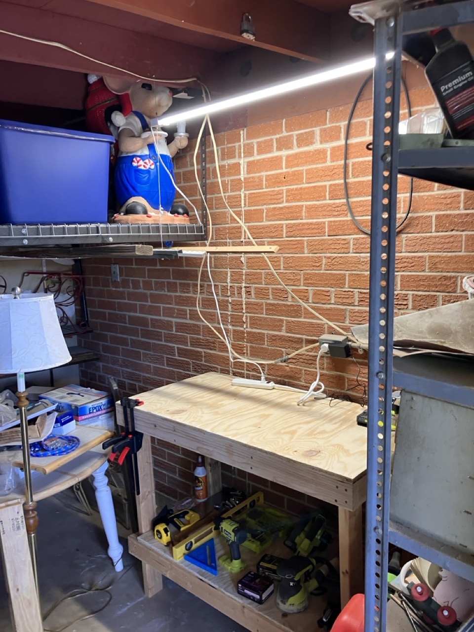 An image taken at a three-quarter angle of workbench against a brick wall. The room is evidently used for storage and is very messy. Hanging from the exposed beams of the room is a 4-foot shop light with wires hanging down. The bench has a lower level filled with various tools and fasteners.