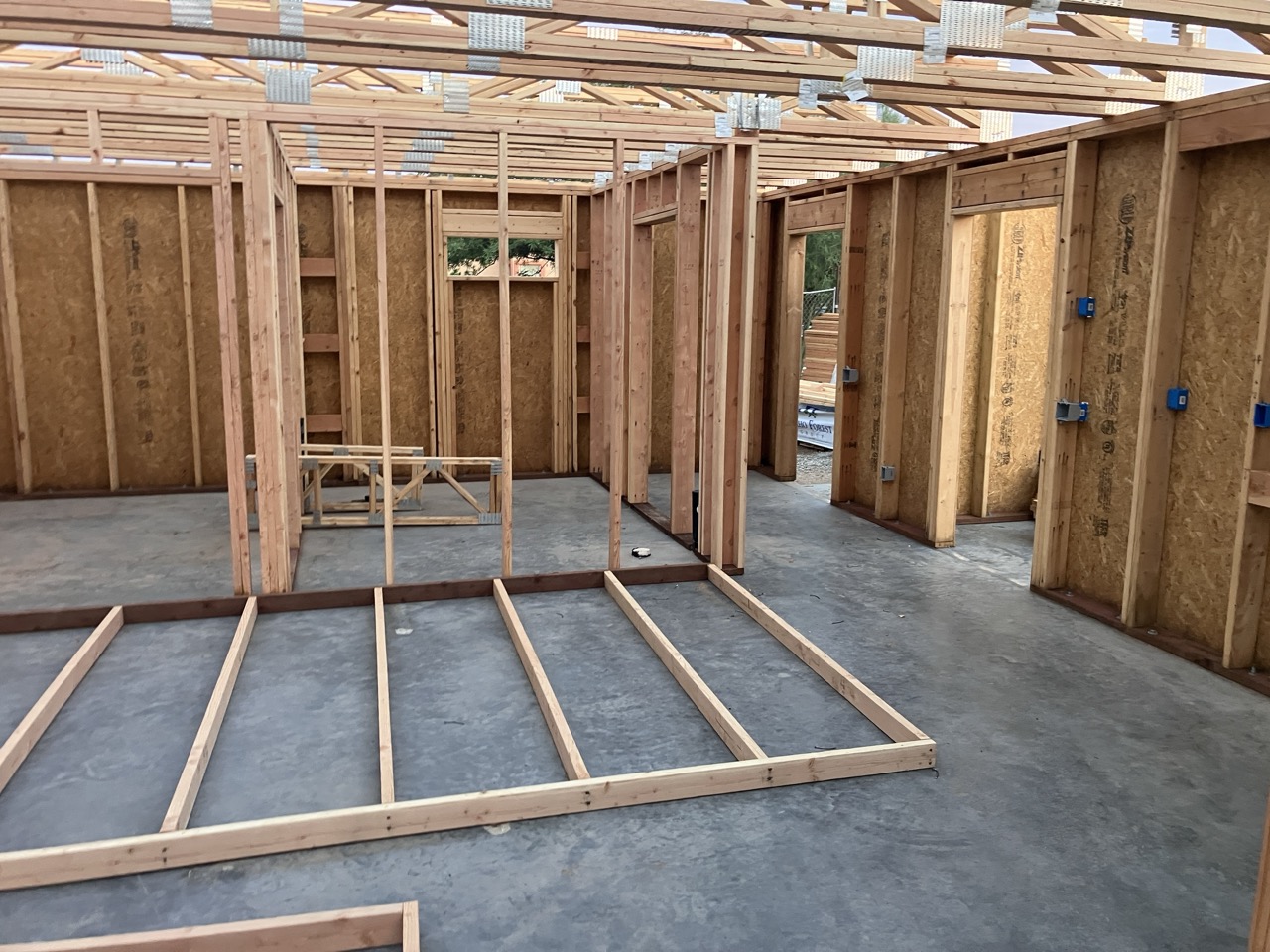 A photo taken of the inside an in-construction house. The walls are completely unfinished and without any drywall. Sitting on the concrete floor is a frame waiting to be lifted up and put into place.