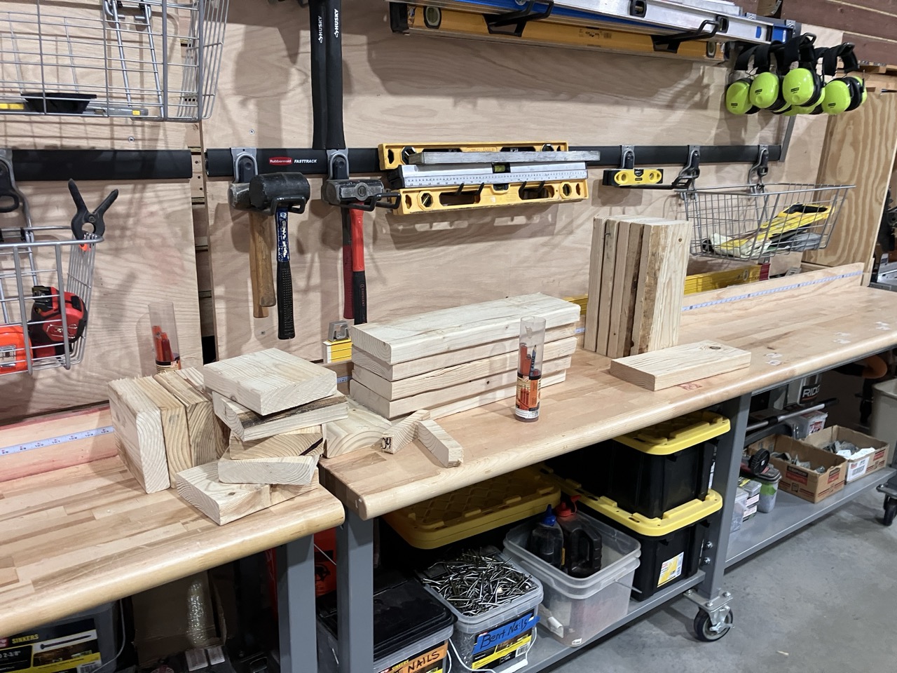 A photo showcasing 2x6 wood boards cut at varying lengths between half a foot and two feet long. The boards are resting in different orientations on a very wide workbench countertop. Mallets, sledgehammers, and measuring tools can be seen around the work space.